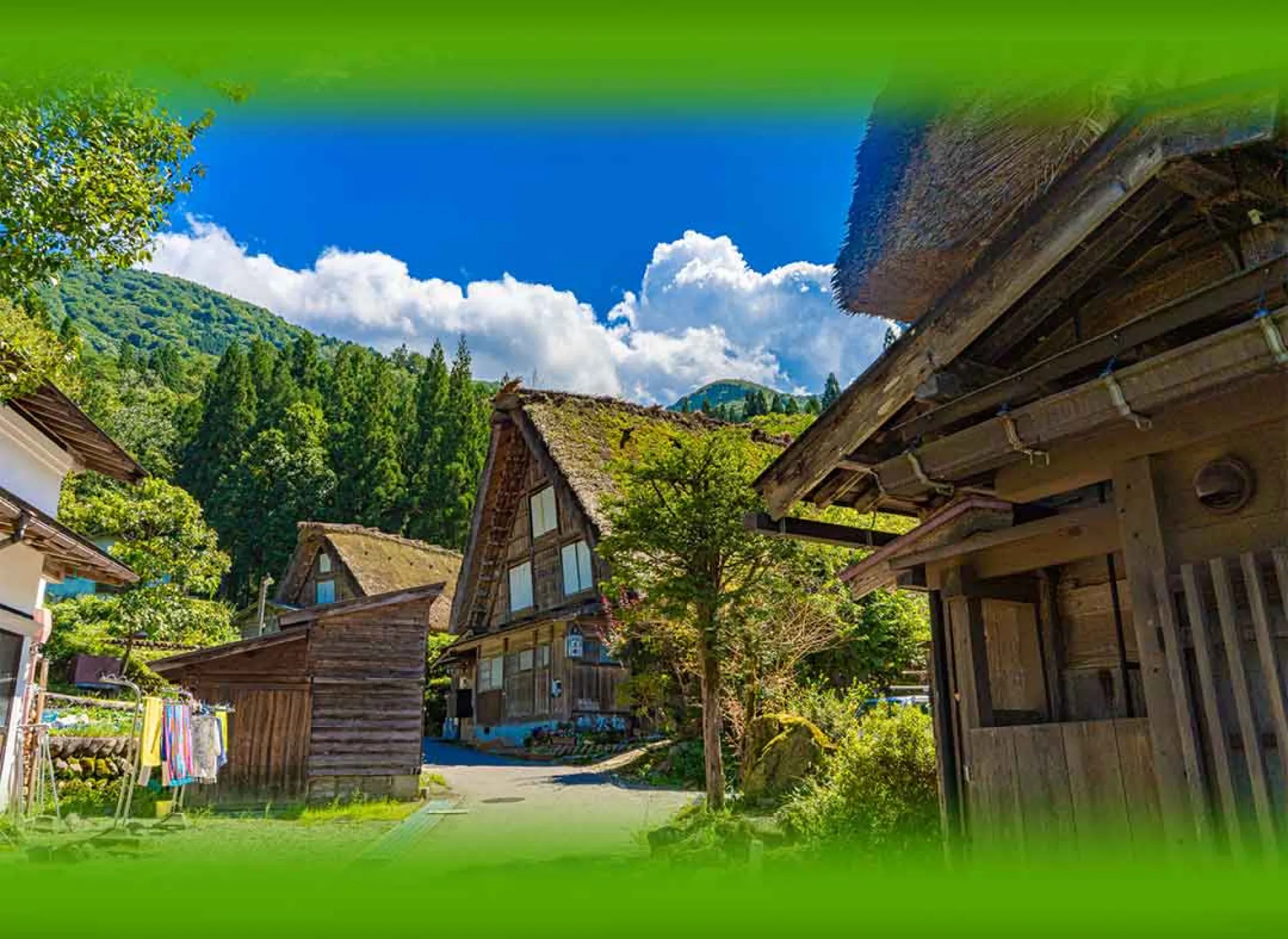 青空と豊かな山々を背景に、青々とした苔がのった伝統的な茅葺き屋根の古民家が並ぶ集落の風景