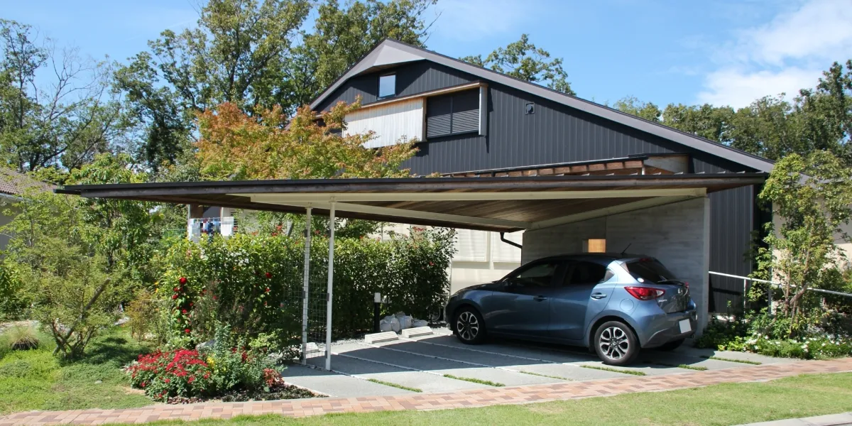 緑豊かな庭園に囲まれた、カーポートに青い車が停まる黒い外壁のモダンな2階建て住宅